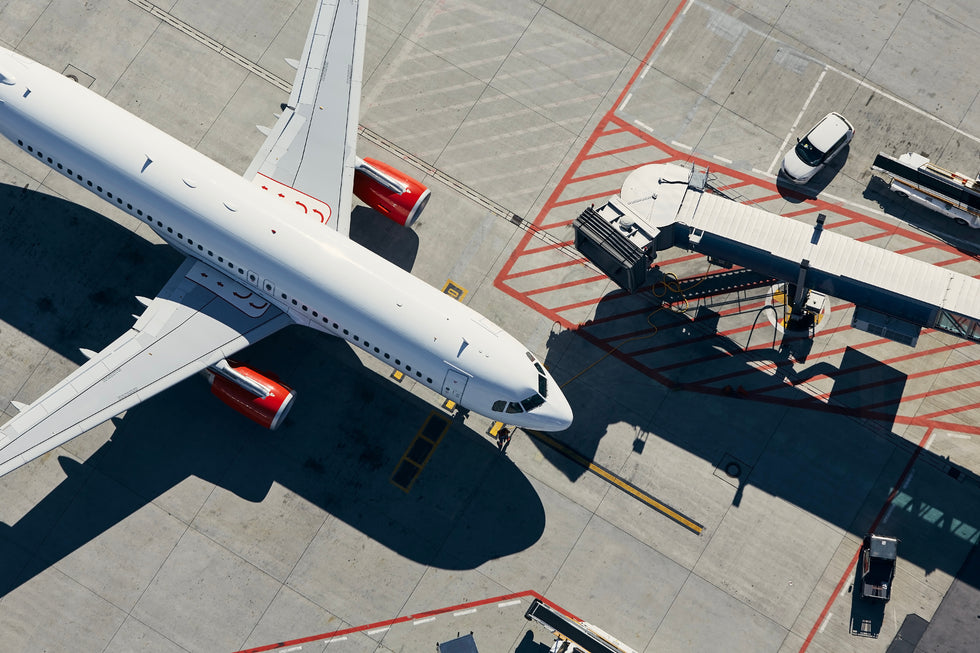 Aerial view of a white commercial airplane parked at an airport gate, with ground vehicles and tarmac markings visible.