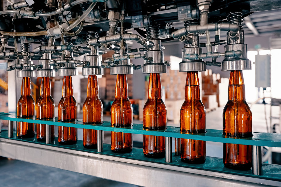 Amber glass beer bottles moving along a conveyor belt in a beverage bottling facility under industrial machinery.