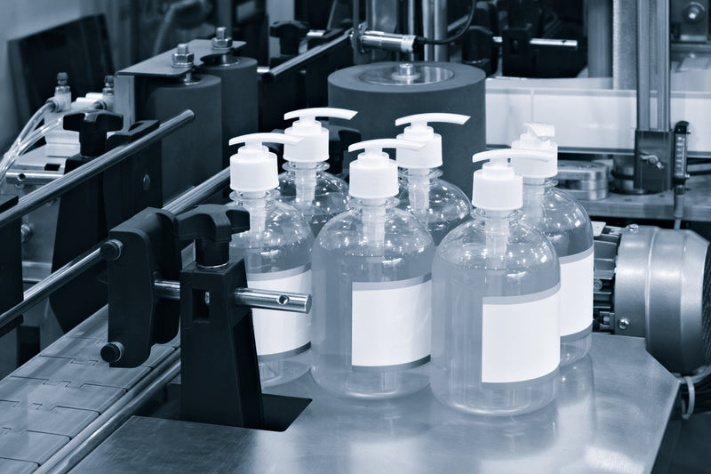 A group of clear plastic pump bottles with blank white labels and white pump dispensers, arranged on a conveyor belt in an industrial setting.