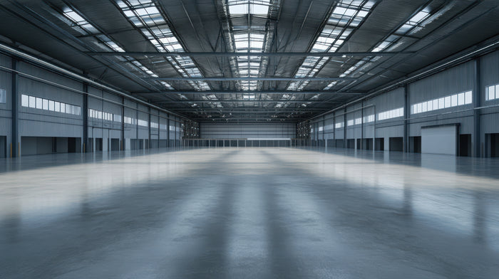 A wide, empty industrial warehouse interior with a polished concrete floor, tall metal support columns, and a high ceiling with skylight panels.