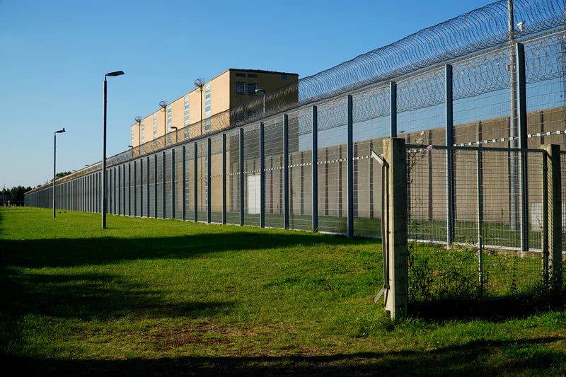 A long metal perimeter fence with barbed wire tops runs alongside a grassy field and a beige building under a clear blue sky.