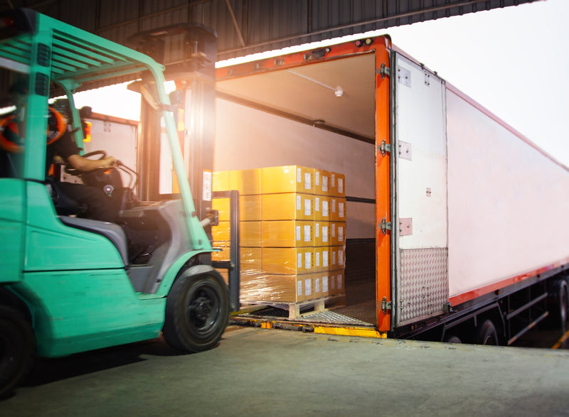A forklift loads yellow stacked boxes onto a covered truck trailer at a warehouse dock.