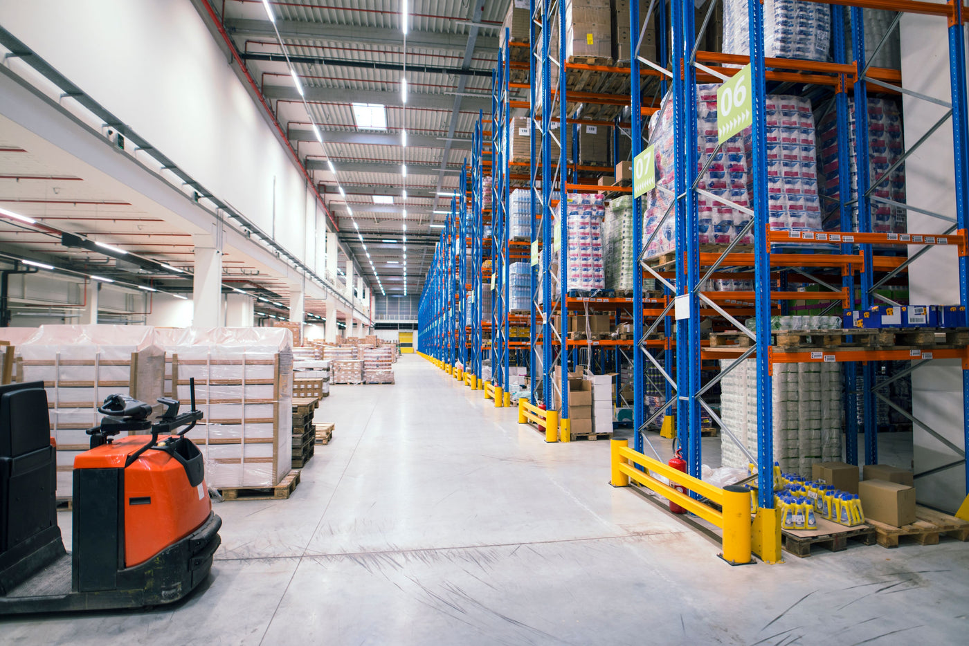 A wide aisle in a large warehouse with tall blue and orange industrial shelving filled with pallets of packaged goods, a concrete floor, overhead lighting, and an orange electric pallet truck in the foreground.