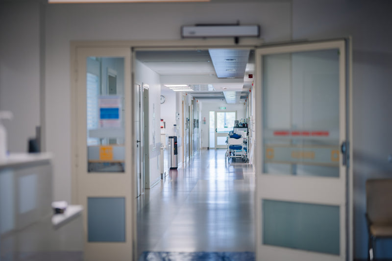 A view through open hospital-style doors into a long, softly blurred hallway with glass doors, fluorescent lighting, and a gurney in the distance.