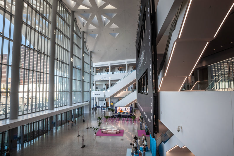 A high-angle view of a modern multi-story building lobby with tall floor-to-ceiling windows, escalators, and a bright open interior space.