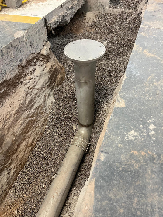 A galvanized steel floor clean out with a circular top cover, installed in a trench filled with gravel next to concrete structures.