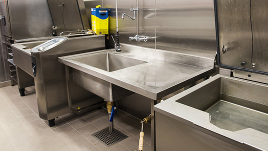 Commercial stainless steel kitchen sinks with integrated faucets and floor sink in a clean industrial kitchen setting.