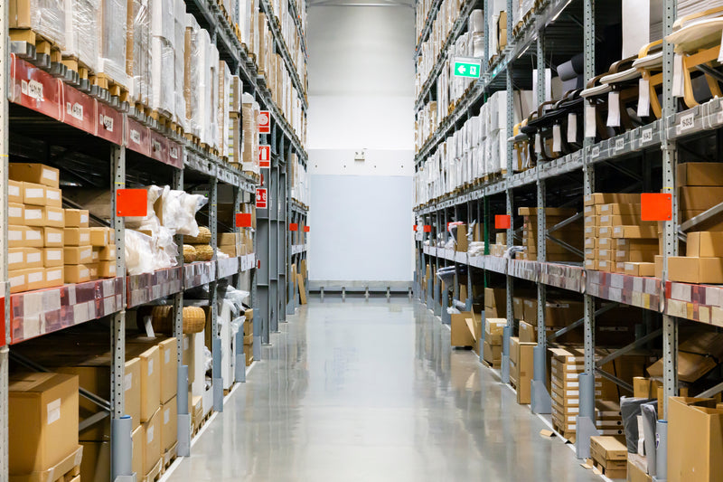 Warehouse aisle with tall racks on each side containing many cardboard boxes and various products.