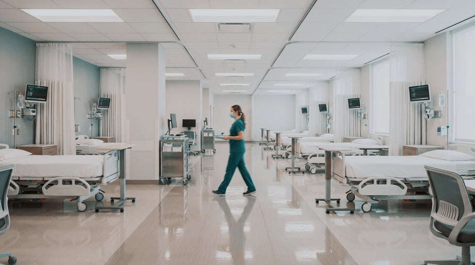 A healthcare worker in teal scrubs walks down a long, bright hospital ward with multiple patient beds, medical carts, and ceiling lights.