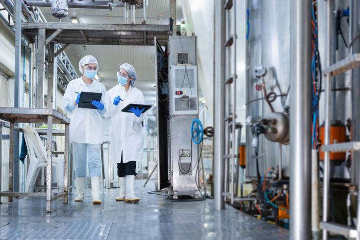 Two scientists in white lab coats, face masks, and hairnets stand in a clean pharmaceutical or biotech production facility, holding tablets to review data. Large stainless steel reactors and industrial equipment line the background of the bright, sterile room.