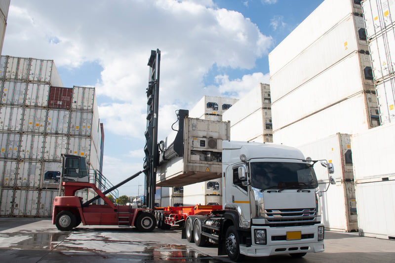 A white semi-truck parked at a docking terminal next to a red container forklift, with stacked shipping containers under a blue sky.