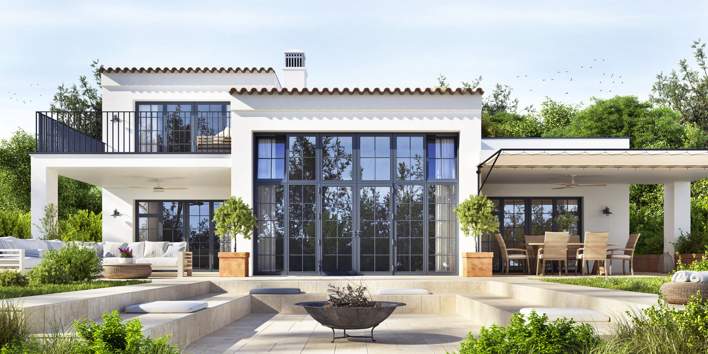 Modern white two-story house with large black-framed glass windows and doors, a covered patio with dining furniture, landscaped greenery, and a light stone courtyard in front.