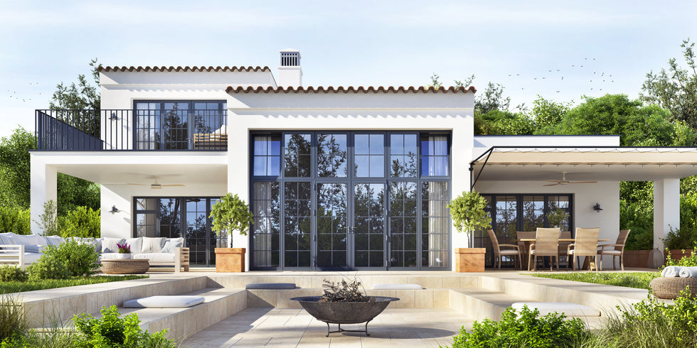 Modern white two-story house with large black-framed glass windows and doors, a covered patio with dining furniture, landscaped greenery, and a light stone courtyard in front.