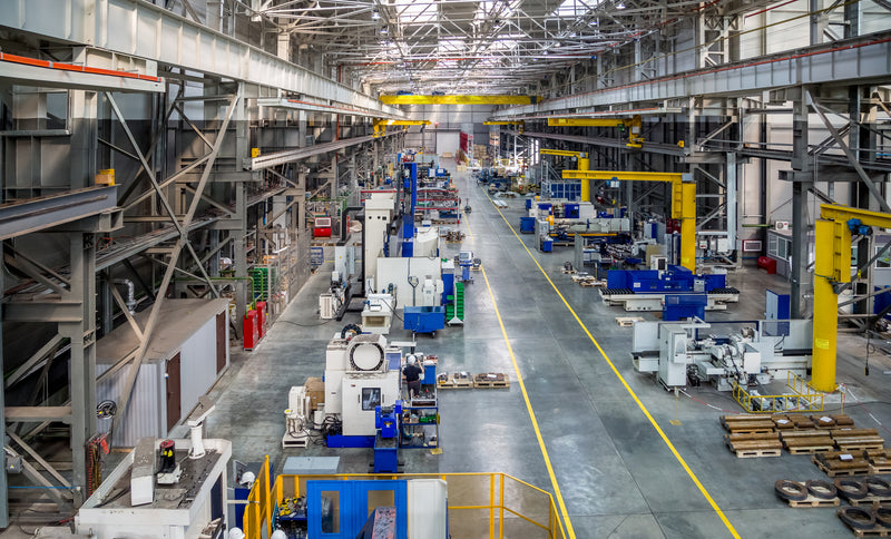 A wide interior view of a large industrial factory floor with rows of blue and white machinery, yellow overhead cranes, metal support structures, and a long concrete aisle.