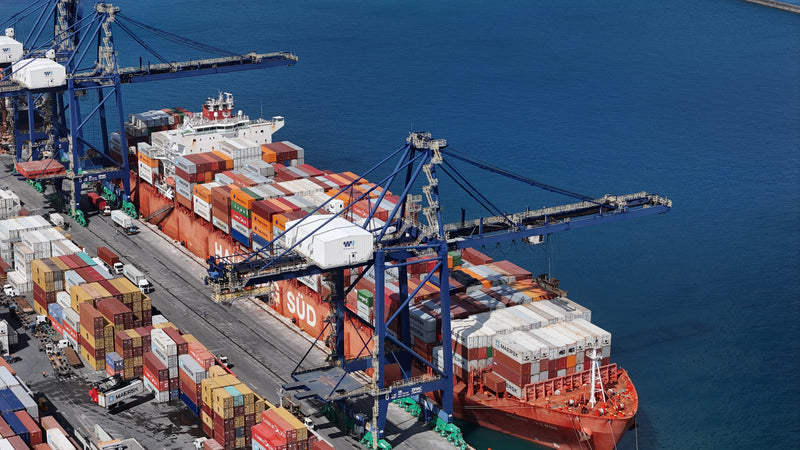Aerial view of a large cargo ship docked at a port with blue cranes, stacked multicolored shipping containers, and blue water surrounding the vessel.