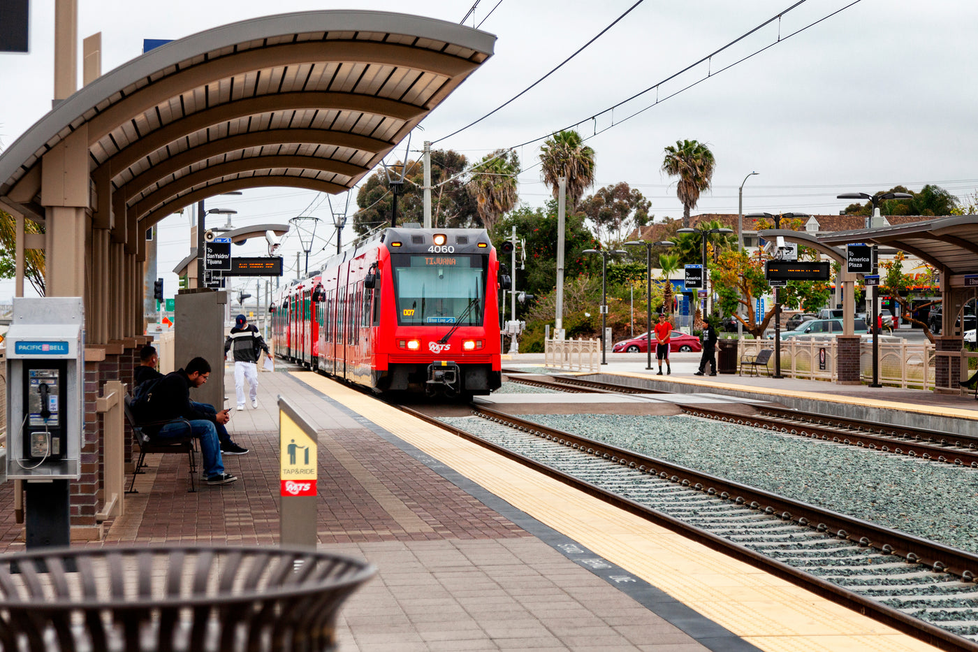 A red train approaches a station where people sit on a platform under a curved roof, with palm trees, and overcast sky in the background.
