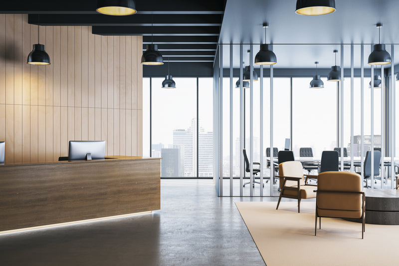 An office lobby with a wood reception desk, black pendant lights, floor-to-ceiling windows, and seating areas with tan chairs and a beige rug.