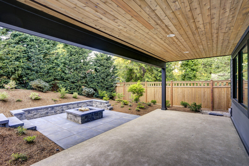 Covered cement patio with stone fireplace with woodchip landscaping. 