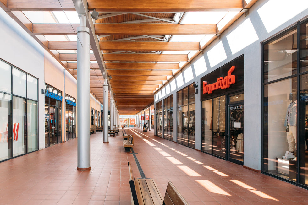 A long shopping mall with storefronts on both sides, glass windows, wooden ceiling beams, and tiled flooring. Store signage and mannequins feature prominently in the windows.