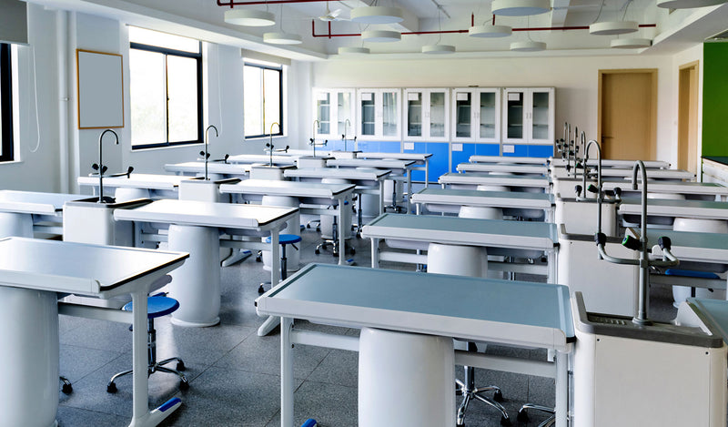A bright, modern science laboratory with multiple white and light blue lab tables, sink stations, chairs, and storage cabinets along the back wall.