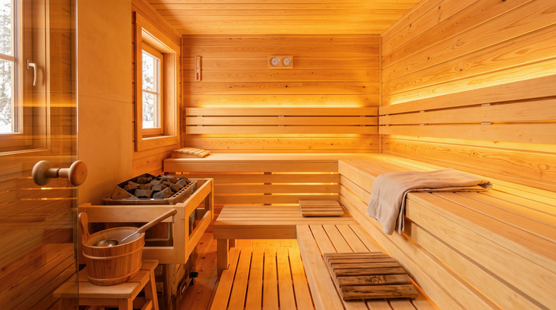A warmly lit wooden sauna interior with tiered seating, a glass door, a pile of heated stones, and a wooden bucket with a ladle in the foreground.