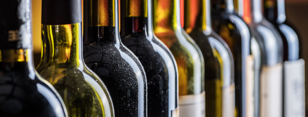 A close-up row of wine bottles with light reflecting through dark glass producing gold and green tones, shown in a shallow depth of field with the front bottle in sharp focus.