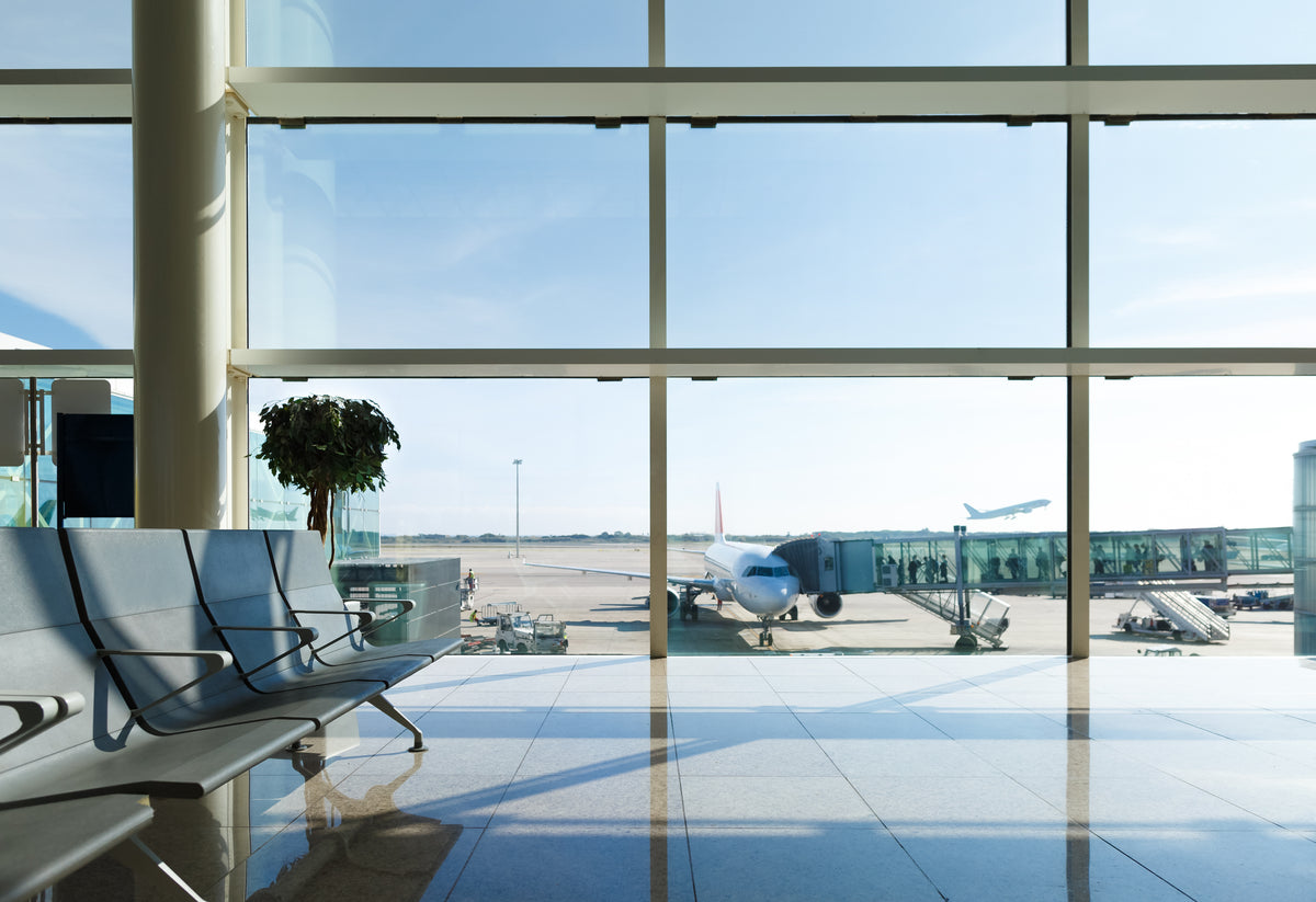 A bright airport terminal interior with large floor-to-ceiling windows, chairs in the foreground, and an airplane visible outside on the tarmac under a clear blue sky.