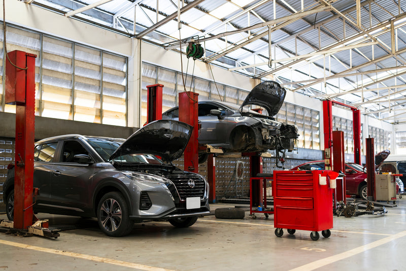 A dark gray SUV with its hood open is parked in a large auto repair garage, alongside a red tool cart and another vehicle raised on a lift. The garage features high ceilings with skylights, red hydraulic lifts, and a concrete floor.