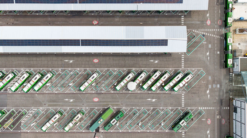 Aerial overhead view of a bus depot with multiple green and white buses parked in designated lanes beside a large covered waiting area with solar panels.