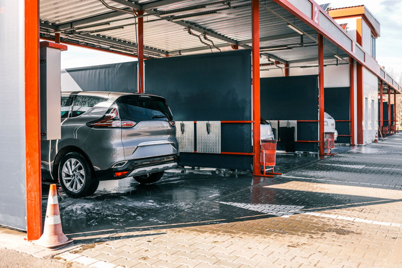 A gray SUV parked under a covered car wash bay with orange support posts, wet pavement, and a traffic cone in the foreground.