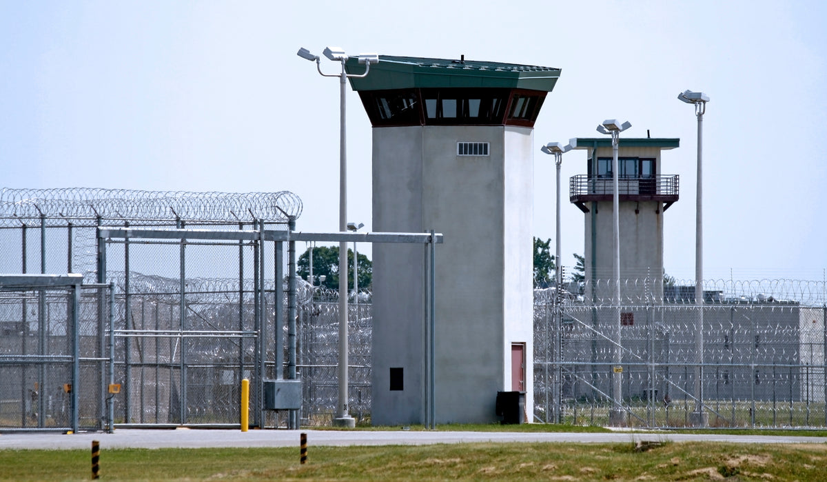 A large prison or detention facility with two tall guard towers, razor wire fencing, and tall light poles under a pale blue sky.