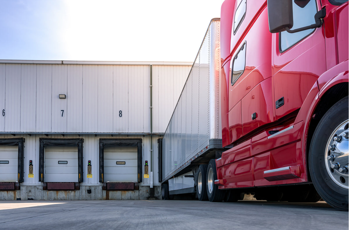 A red semi-truck cab and trailer parked at a loading dock next to a white warehouse building under a bright sky.