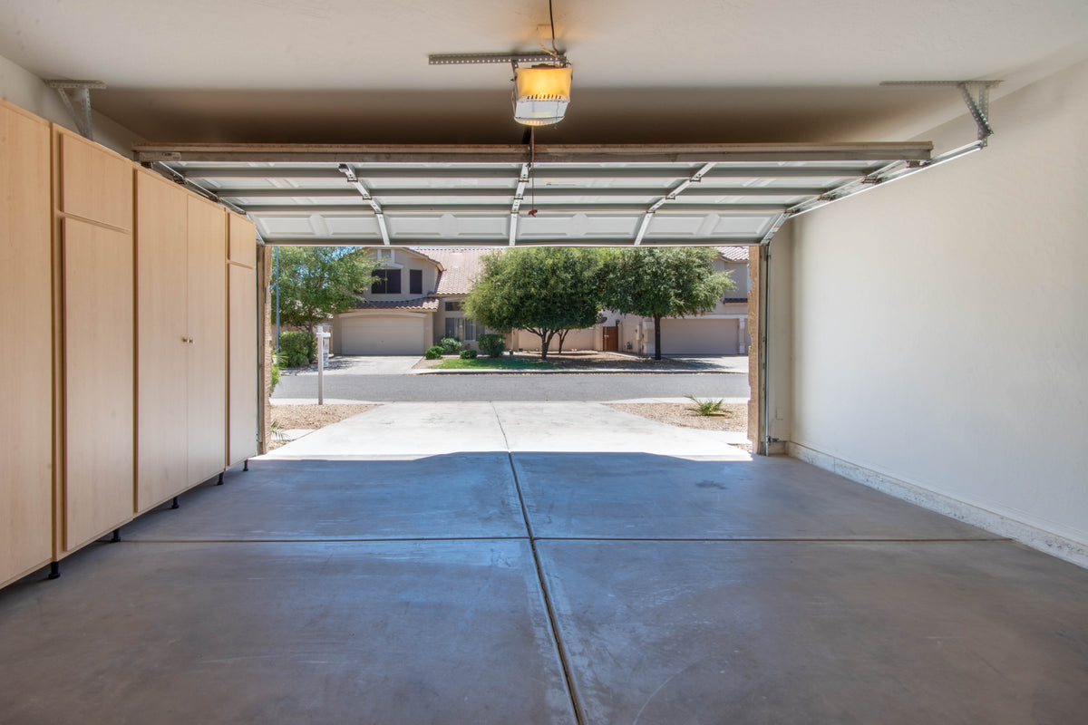 An empty garage interior with a concrete floor, beige walls, a metal overhead door track and panels, and a light fixture. Through the open garage door, a suburban street with trees and houses is visible.