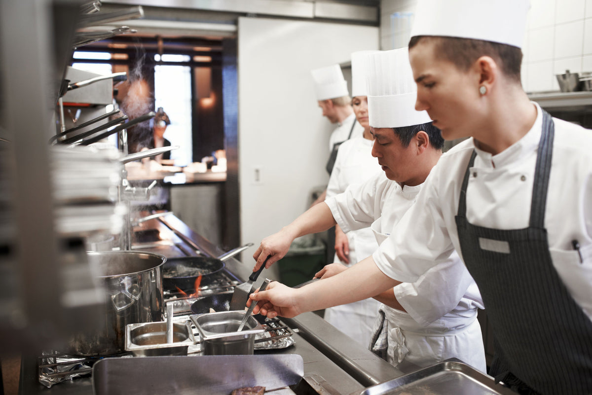 A group of professional chefs in white uniforms and tall white hats working at a stainless steel kitchen station, with one chef in the foreground wearing a dark apron.