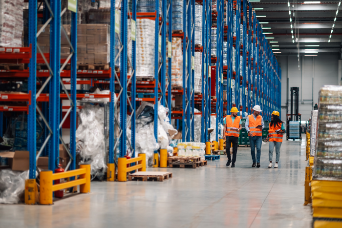 Three workers in high-visibility safety vests and hard hats walk through a large, brightly lit warehouse aisle lined with blue and red industrial shelving units.