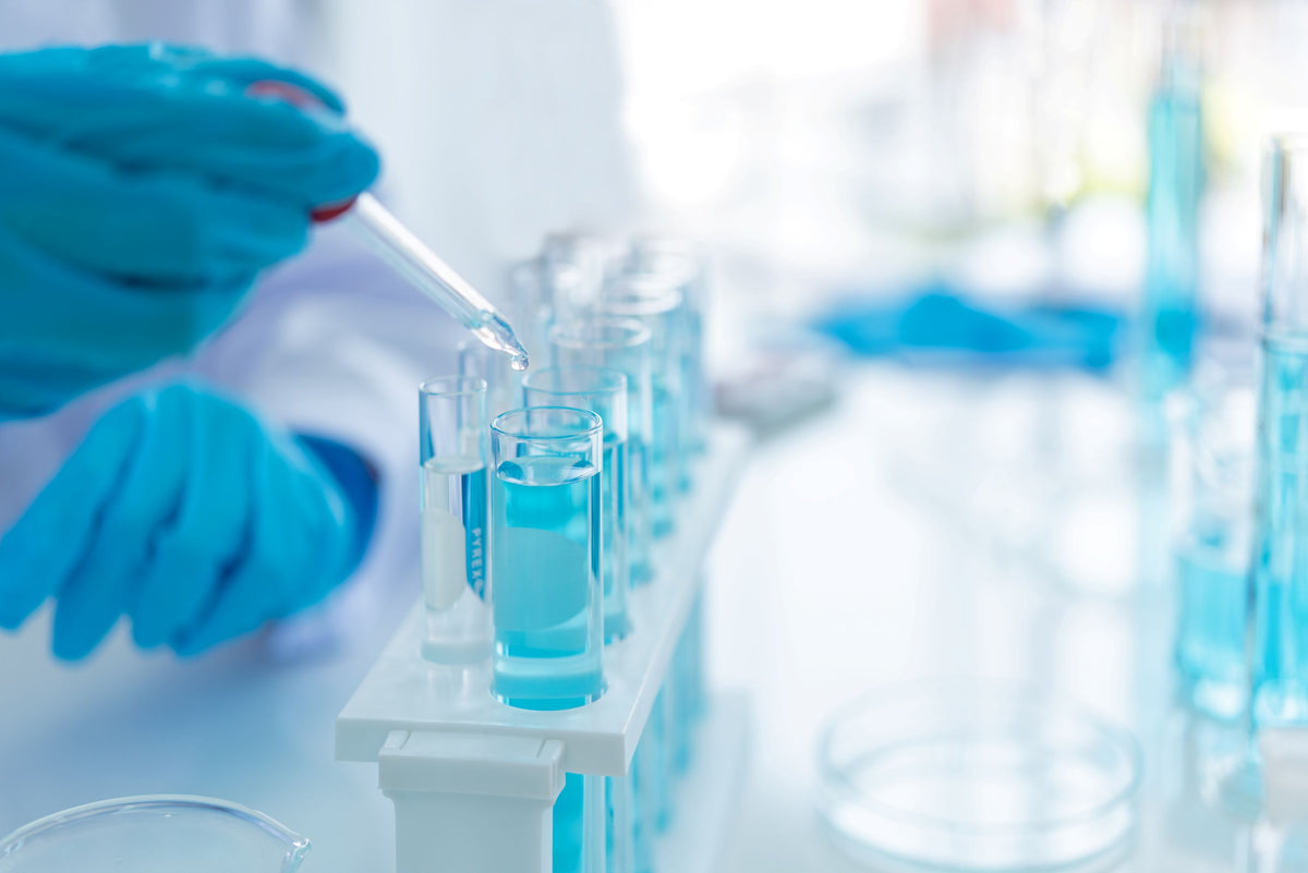 A close-up of a person wearing blue gloves using a pipette to transfer liquid into a test tube rack, with blue liquid visible in several tubes in a bright laboratory setting.